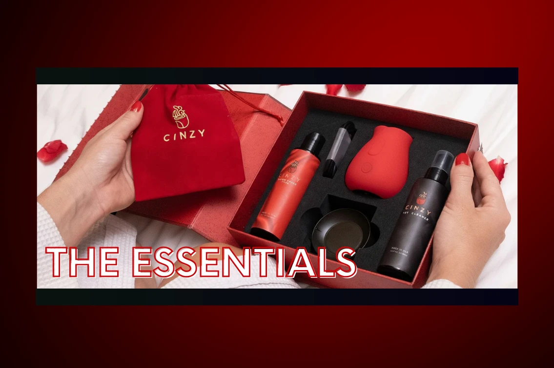 Hands holding open the Cinzy luxury gift box on white bedding with rose petals. The essentials include the red rose toy, water-based lube, toy cleaner, charging base, drawstring pouch, and instruction manual.