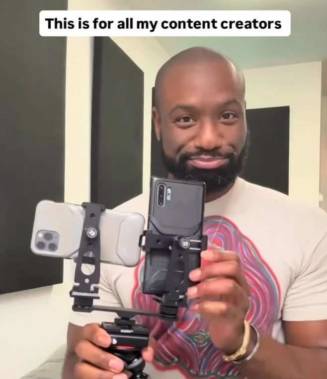 A smiling man with a beard wearing a white t-shirt holds up a black dual-phone rig on a tripod. The rig has two smartphones mounted side-by-side — one light-colored iPhone and one dark Android phone — facing forward. A text overlay at the top reads: 'This is for all my content creators'. He is looking directly at the camera with a friendly expression.