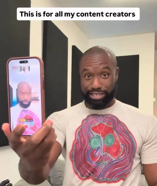 A man with a beard and white graphic t-shirt holds up a smartphone in a pink case toward the camera. The phone screen shows a live recording or video call of himself. He has wide eyes and an excited expression as he speaks. A text overlay at the top reads: 'This is for all my content creators'. The background shows a room with black acoustic panels.